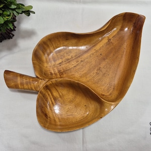 Puede incluir: Bandeja de madera para servir con forma de hoja. La bandeja es de madera de color marrón claro y tiene un asa. Tiene dos secciones para servir alimentos o exhibir artículos. La veta de la madera es visible.