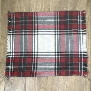 May include: A rectangular fabric throw blanket featuring a classic plaid pattern in shades of deep red, charcoal gray, black, and white. The textile has a soft, woven texture and is finished with frayed, fringed edges along all four sides.