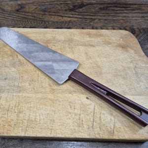 May include: A cake server with a stainless steel blade and a dark brown handle. The server is on a well-used wooden chopping board. The handle has a distinctive, open design.
