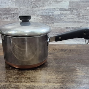 May include: Stainless steel saucepan with a lid and a black handle. The pot has a copper-colored bottom and a silver-colored lid with a black knob. The handle has a metal loop at the end.