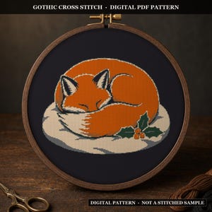 Pode incluir: Um bastidor de bordado circular de madeira contendo um padrão de ponto cruz de uma raposa laranja adormecida enrolada numa mancha de neve branca com folhas de azevinho verdes. Texto GOTHIC CROSS STITCH DIGITAL PDF PATTERN.