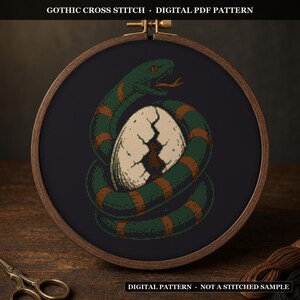May include: A circular wooden embroidery hoop containing a cross stitch pattern on black fabric. The design features a green and orange snake coiled around a large, cracked white egg. Text at the top reads GOTHIC CROSS STITCH DIGITAL PDF PATTERN.