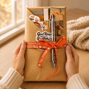 May include: A brown paper envelope containing stationery items, including a pen with a metallic finish, stickers, and cards. The envelope is tied with an orange ribbon featuring the text HELLO FALL and a sticker reading Good Things happen after coffee.