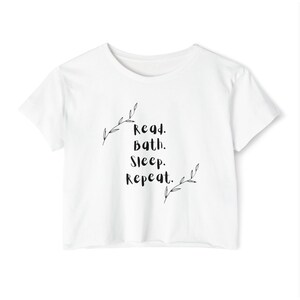 May include: A white short-sleeved t-shirt featuring black text that reads Read. Bath. Sleep. Repeat. The text is flanked by two delicate, hand-drawn style black leaf sprigs, one positioned at the top left and one at the bottom right of the lettering.