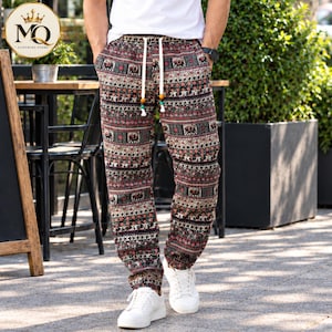 May include: Loose-fitting trousers featuring a multi-colored horizontal pattern with repeating elephant motifs and geometric shapes. The pants include a white drawstring waistband with decorative beads at the ends and are paired with white sneakers.