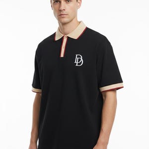 Könnte beinhalten: Ein schwarzes kurzärmeliges Poloshirt mit beigem Kragen und Ärmelbündchen, die einen dünnen roten Rand aufweisen. Das Shirt verfügt über eine Knopfleiste mit beigen und roten Details sowie ein weißes, gesticktes D-Logo auf der linken Brust.