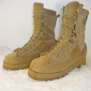 Könnte beinhalten: Ein Paar taktische Stiefel aus beigem Wildleder und Canvas mit hohem Schaft, verstärkter runder Zehenkappe und passenden beigen Schnürsenkeln, die durch Metallösen und Haken geführt werden. Die Stiefel haben dicke, profilierte Gummisohlen.