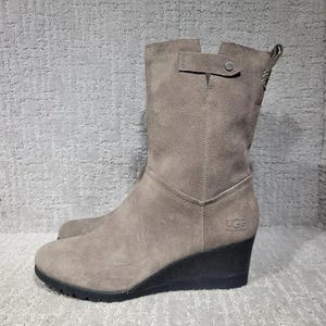以下が含まれることがあります： トープ色のスエード素材のミッドカーフウェッジブーツ。黒いラバー製のウェッジソール、小さな金属製ボタンが付いたサイドストラップ、外側下部に刻印されたUGGのロゴが特徴です。丸みを帯びたトゥと、すっきりとした構造的なシルエットが魅力です。