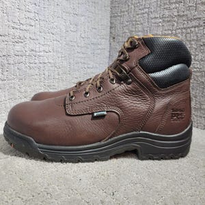 Puede incluir: Una bota de trabajo de cuero marrón con cuello acolchado negro, cordones marrón oscuro con ojales metálicos y una suela de goma negra gruesa. El lateral muestra una etiqueta impermeable y el logotipo Timberland PRO grabado en el talón.