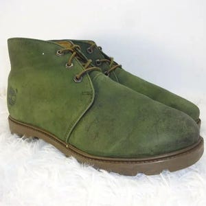 May include: A pair of olive green ankle boots featuring a smooth suede-like finish, brown laces, metal eyelets, and a thick brown rubber sole. The side of the boot displays a small embossed tree logo, and the design includes a rounded toe box.