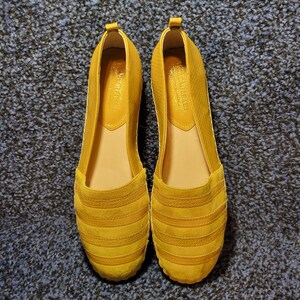 May include: A pair of mustard yellow slip-on flats featuring a textured, multi-striped upper design. The shoes have a rounded toe, a low-cut opening, and a smooth interior lining with a visible brand logo printed on the heel area of the insole.