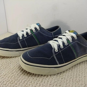 May include: A pair of low-top navy blue sneakers featuring white laces, a thin green vertical stripe detail on the side, and a thick off-white rubber sole. The shoes have visible white stitching along the panels and a padded collar around the ankle.