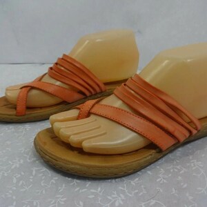 May include: A pair of orange sandals featuring multiple thin straps across the top and a toe loop design. The sandals have a flat, tan-colored sole with a textured edge and are displayed on beige foot-shaped forms to show their structure.