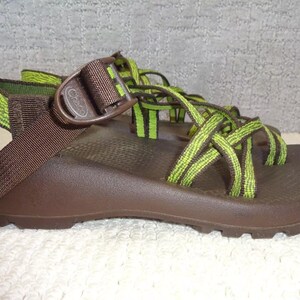 May include: A brown sandal with lime green and dark green patterned webbing straps. The footwear features a brown plastic buckle with the word Chaco embossed on the side, a contoured brown footbed, and a textured sole for traction and support.
