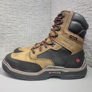 May include: A brown and black leather work boot featuring a high-top design, black padded collar, and red-accented laces. The side displays the text Wolverine, Waterproof Plus, and DuraShocks Flex, with a black rubber toe cap and a thick lugged sole.