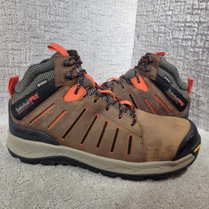 May include: A pair of brown leather Timberland PRO work boots featuring orange accents, black laces, and a black composite safety toe. The boots have a waterproof design, a tan midsole, and a black outsole with the Vibram logo on the side.