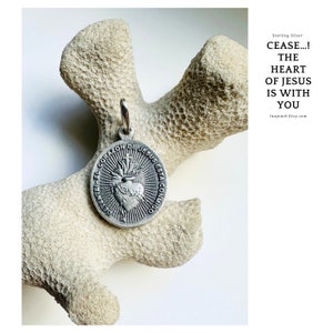 Puede incluir: Un colgante de plata con un diseño de corazón y el texto "SELENTEEL EL CORAZON DE JESUS ESTA CONMIGO". El colgante está sobre una superficie blanca de tipo coral. Plata de ley CESE... EL CORAZÓN DE JESÚS ESTÁ CONTIGO Inajewel.Etsy.com