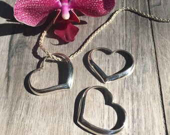 May include: A silver necklace with three heart-shaped pendants. The necklace is lying on a wooden surface with a pink orchid flower.
