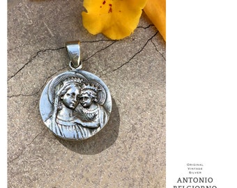 May include: Silver pendant featuring a detailed relief of the Virgin Mary and baby Jesus. The pendant is round and has a halo around the heads of the figures. The pendant is on a textured surface.