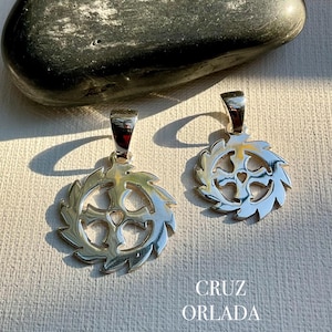 May include: Two silver-colored pendants with a cross design at the center, surrounded by a circular, spiky frame. The pendants are displayed on a textured, light-colored surface. The text "CRUZ ORLADA" is visible below the pendants.