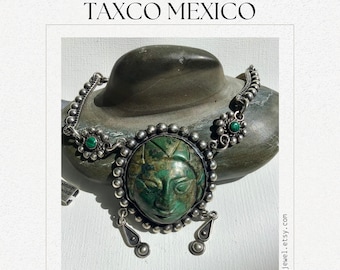 May include: A silver necklace with a green stone pendant featuring a carved face. The pendant is surrounded by silver beads and the necklace has a silver chain with green stone accents. "MID-CENTURY TAXCO MEXICO" is written in the image.