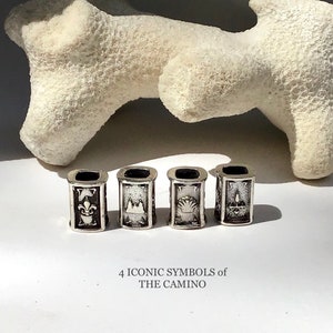 Puede incluir: Cuatro cuentas de plata con diferentes símbolos, incluyendo una flor de lis, una concha de vieira, una iglesia y una vela. El texto "4 ICONIC SYMBOLS of THE CAMINO" está debajo de las cuentas.
