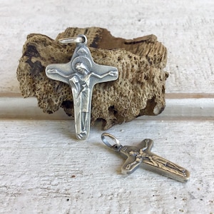 May include: Two silver crucifix pendants, one larger than the other, with detailed carvings of Jesus Christ on the cross. The larger pendant is resting on a piece of driftwood.