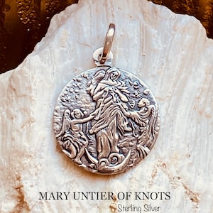May include: A silver pendant featuring a depiction of Mary, the Untier of Knots, with a detailed design of angels and knots. The pendant is labeled "Mary Untier of Knots Sterling Silver".
