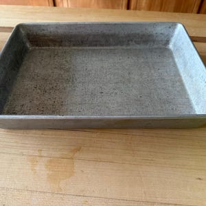 May include: A rectangular metal baking tray with a silver-toned finish and a flat base. The interior surface of the tray shows visible signs of wear, including light scratches and scuffs across the bottom, with straight, raised vertical edges.