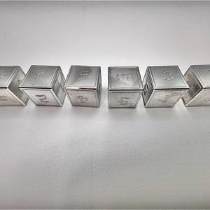 Könnte beinhalten: Ein Set aus sechs silberfarbenen Metallwürfeln, die in einer horizontalen Reihe angeordnet sind. Jeder Würfel zeigt auf seiner Oberseite eingravierte Zahlen in einer stilisierten gotischen Schrift. Die Oberflächen haben eine gebürstete Textur.