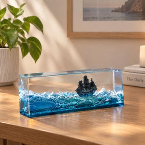 Può includere: Un blocco rettangolare in resina trasparente contenente un modello in miniatura di una nave a vela nera, inserito in una scena di onde oceaniche scolpite in blu e bianco. L'oggetto decorativo mostra la nave che naviga su acqua testurizzata e spumosa.