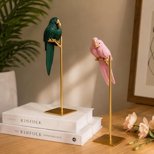 May include: Two decorative parrot figurines perched on thin gold metal stands with square bases. One parrot is dark green with a gold beak, and the other is light pink with a gold beak. The figures are positioned on top of two stacked white books.