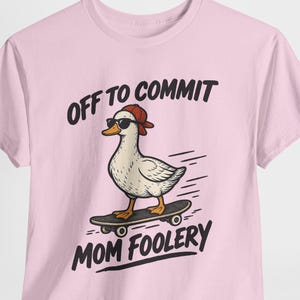 May include: A light pink t-shirt featuring a graphic of a white duck wearing a red baseball cap and black sunglasses while riding a skateboard. The text above and below the duck reads OFF TO COMMIT MOM FOOLERY in bold black capital letters.