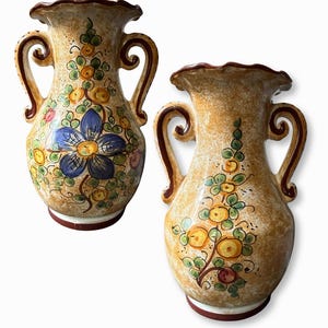 May include: Two ceramic vases with two handles each, featuring a mottled tan and beige glaze. The vases are decorated with hand-painted floral motifs in blue, yellow, and green, and have a scalloped rim with a dark brown painted edge.