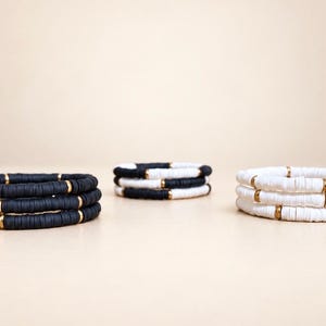 May include: Three stacks of bracelets featuring flat circular beads. The left stack is solid black, the right stack is solid white, and the center stack alternates between black and white beads. Each bracelet includes small gold-toned metal spacers.