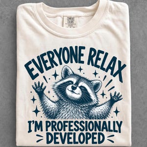 May include: A cream-colored t-shirt featuring a dark blue graphic of a raccoon with its arms raised. The text above reads EVERYONE RELAX and the text below reads I'M PROFESSIONALLY DEVELOPED. The shirt has a Comfort Colors brand label.