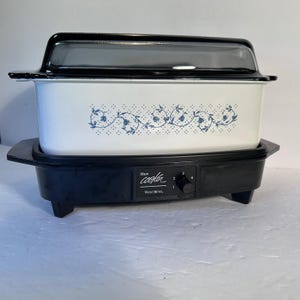May include: A rectangular West Bend slow cooker with a white metal insert featuring a blue floral vine pattern. The appliance has a black base with a control knob numbered one through four and a black lid with a clear glass centre panel.