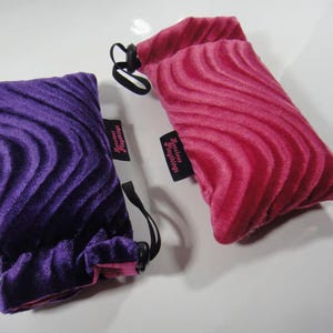 May include: Two velvet drawstring pouches with a wavy pattern. The pouch on the left is purple and the pouch on the right is pink. Both pouches have a small tag that says "Secret Squirrel Things".