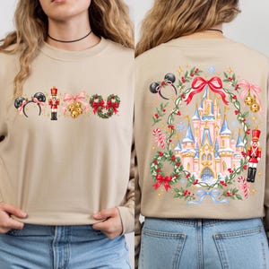 May include: A beige long-sleeve sweatshirt featuring a front graphic of mouse ears, a nutcracker, gold bells, and a wreath. The back graphic displays a large castle surrounded by a holiday wreath, a nutcracker, candy canes, and bows.