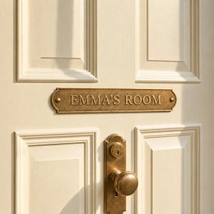 Puede incluir: Una placa de puerta rectangular de latón con extremos redondeados, que presenta el texto grabado EMMA'S ROOM en letras mayúsculas. La placa está montada en una puerta de paneles de color crema sobre un pomo y cerradura de latón a juego.