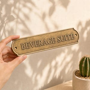 Puede incluir: Un letrero rectangular de metal en tono lat&oacute;n con esquinas redondeadas y bordes decorativos, que presenta el texto en relieve BEVERAGE SUITE en may&uacute;sculas. El letrero incluye dos peque&ntilde;os orificios circulares de montaje a los lados.
