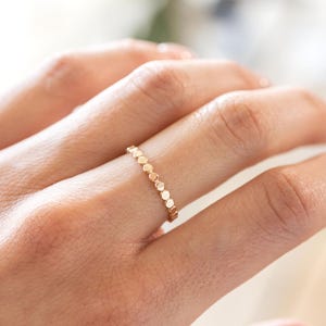 Puede incluir: Un delicado anillo de oro con una serie de pequeños acentos circulares espaciados uniformemente. El anillo se lleva en un dedo, mostrando su diseño simple pero elegante. El color dorado cálido del anillo contrasta con el tono de la piel.