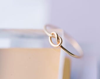 Puede incluir: Un delicado brazalete dorado con un diseño de nudo. El brazalete es delgado y parece estar hecho de metal. El fondo está borroso, con una paleta de colores suaves y neutros.