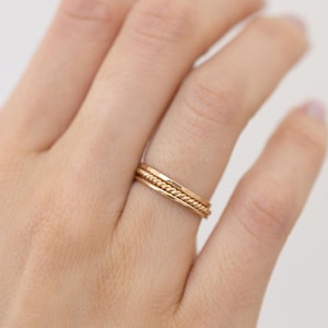 May include: Three gold rings stacked on a finger. The rings have different textures: one is smooth, one has a hammered finish, and the third has a twisted rope design. The rings are a warm gold color.