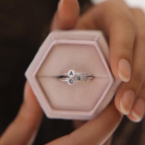 May include: Three silver rings, each with a small, circular charm bearing the letters A, B, and C. The rings are presented in a pink, hexagonal jewellery box. The rings are held by a person.