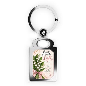 May include: A silver-toned metal keychain featuring a rectangular pendant with a printed design of white lily of the valley flowers tied with a pink ribbon. The text reads Little Light, Bright Care Kinder Days, You Make A Difference.