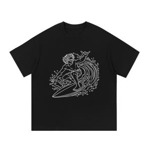 Puede incluir: Una camiseta negra de manga corta con un gráfico de líneas blancas que muestra a una persona surfeando una ola. El surfista lleva gafas de sol y hace un gesto con la mano mientras monta sobre una tabla de surf entre salpicaduras de agua.