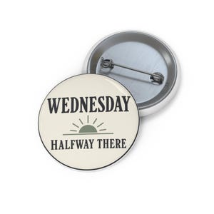 Op de afbeelding: Een ronde pin-back button met een crèmekleurige voorzijde met de zwarte tekst WEDNESDAY en HALFWAY THERE, gescheiden door een groen halfrond zon-icoon. De achterkant van de button is van zilverkleurig metaal met een horizontale speld.