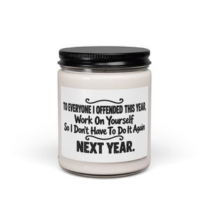 Puede incluir: Una vela de cera blanca en un frasco de vidrio transparente con tapa de metal negro. Una etiqueta rectangular blanca en la parte frontal presenta el texto en negro: TO EVERYONE I OFFENDED THIS YEAR, Work On Yourself So I Don't Have To Do It Again NEXT YEAR.