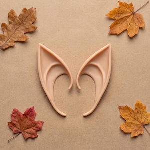 May include: A pair of light beige, pointed prosthetic ears with a smooth, curved design. The ears feature a tapered, elongated shape with a defined inner ridge and a curved base, designed to mimic a fantasy creature aesthetic for costume use.